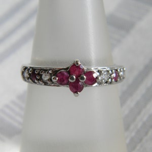May include: A silver ring with a flower design featuring five red gemstones and smaller white gemstones around the band.