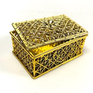 May include: A gold-colored decorative box with an open lid. The box features an intricate lattice design and is adorned with several clear, sparkling embellishments. The box is rectangular and sits on a white surface.