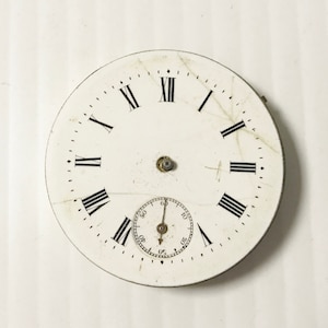 Puede incluir: Esfera de reloj de bolsillo blanca con marcadores de hora en n&uacute;meros romanos y un c&iacute;rculo conc&eacute;ntrico m&aacute;s peque&ntilde;o con marcas negras para los segundos. La esfera del reloj tiene un aspecto desgastado y envejecido.