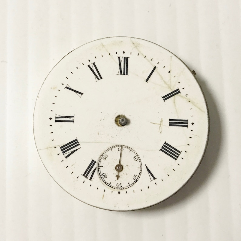 Puede incluir: Esfera de reloj de bolsillo blanca con marcadores de hora en n&uacute;meros romanos y un c&iacute;rculo conc&eacute;ntrico m&aacute;s peque&ntilde;o con marcas negras para los segundos. La esfera del reloj tiene un aspecto desgastado y envejecido.
