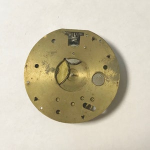 Puede incluir: Un movimiento de reloj de metal de color dorado con engranajes expuestos. El movimiento es circular y tiene un di&aacute;metro de aproximadamente 7,6 cm.