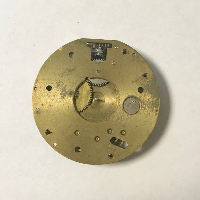 Puede incluir: Un movimiento de reloj de metal de color dorado con engranajes expuestos. El movimiento es circular y tiene un di&aacute;metro de aproximadamente 7,6 cm.