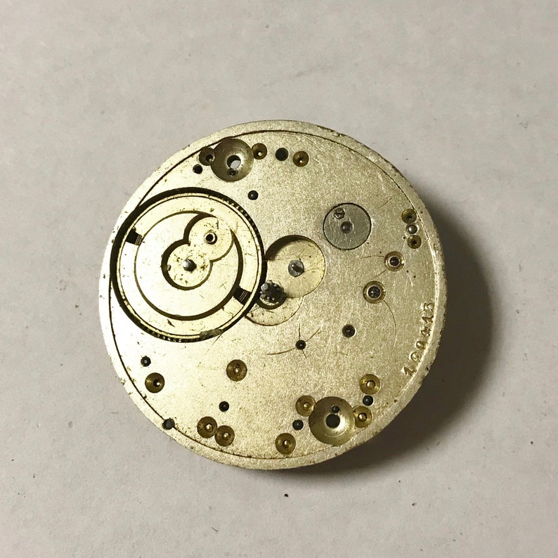 Puede incluir: Primer plano de un movimiento de reloj de color dorado con engranajes intrincados y un dise&ntilde;o circular. El movimiento tiene el n&uacute;mero "17004413" impreso en el lateral.