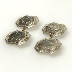 May include: A pair of silver cufflinks with an octagonal design. Each cufflink has a central oval shape and is surrounded by geometric patterns. The cufflinks are connected by a small bar.