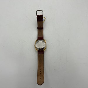 Puede incluir: Un reloj de pulsera dorado con una esfera cuadrada y una correa de cuero marr&oacute;n. El reloj tiene una esfera blanca y un bisel plateado.