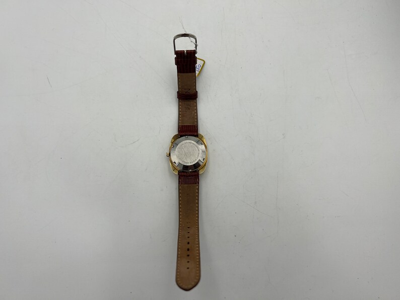 Puede incluir: Un reloj de pulsera dorado con una esfera cuadrada y una correa de cuero marr&oacute;n. El reloj tiene una esfera blanca y un bisel plateado.