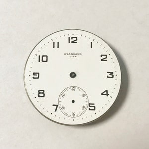 May include: White watch face with black numbers and hands. The watch face has the text "STANDARD U.S.A." printed on it.