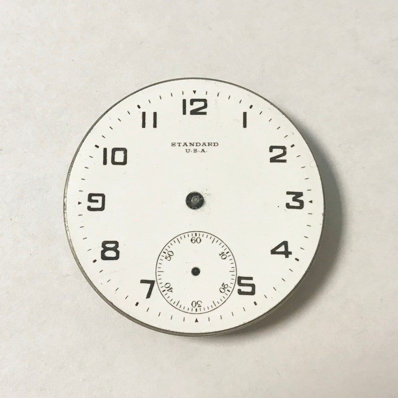 May include: White watch face with black numbers and hands. The watch face has the text "STANDARD U.S.A." printed on it.
