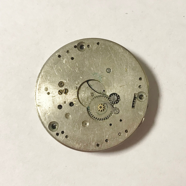 May include: A silver-toned watch movement with visible gears and a circular design. The movement is disassembled and shows the inner workings of the watch.
