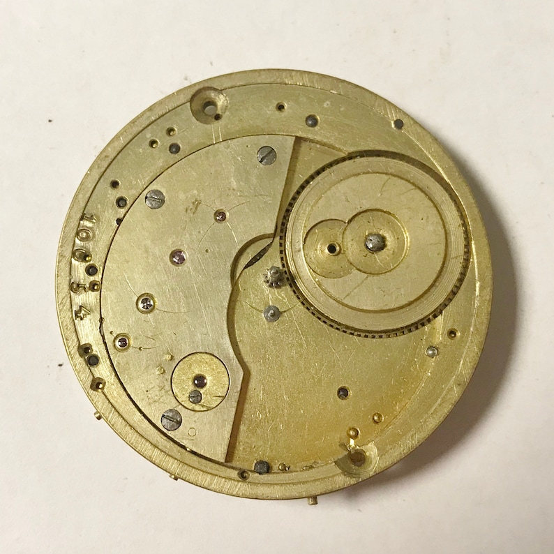 May include: A gold-toned watch movement, showcasing intricate gears, screws, and other small parts. The circular mechanism, likely from a pocket watch, displays a complex arrangement of components. The watch movement is a vintage item.