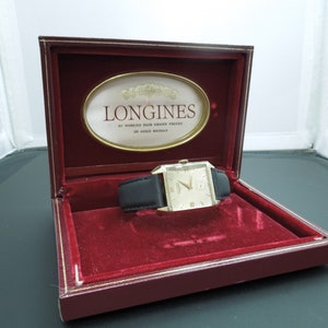 May include: A gold Longines wristwatch with a black leather band in a red velvet lined box. The box has a gold oval label with the text "Longines 20 World's Fair Grand Prizes 28 Gold Medals".