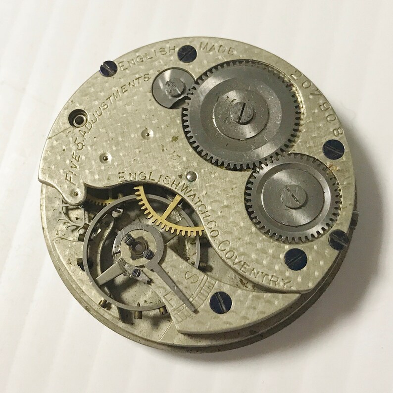 Puede incluir: Primer plano de un movimiento de reloj de bolsillo plateado con la inscripci&oacute;n "English Watch Co. Coventry" y "Five Adjustments English Made 207908".
