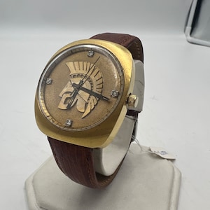 Puede incluir: Un reloj de pulsera dorado con una correa de cuero marr&oacute;n. La esfera del reloj presenta una imagen en relieve de un casco de soldado romano con diamantes alrededor del borde.