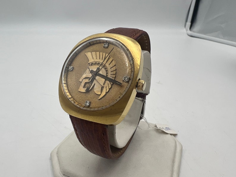 Puede incluir: Un reloj de pulsera dorado con una correa de cuero marr&oacute;n. La esfera del reloj presenta una imagen en relieve de un casco de soldado romano con diamantes alrededor del borde.