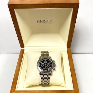 May include: A Zenith watch with a black face and silver band rests on a cream-colored pillow inside a wooden box. The box is open, revealing the watch and the Zenith logo with the text "LIFE IS IN THE MOVEMENT".