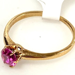 May include: A gold ring featuring a vibrant pink gemstone. The ring's band is a simple, polished gold, and the gemstone is set in a delicate, ornate setting. The ring is a classic piece of jewelry.