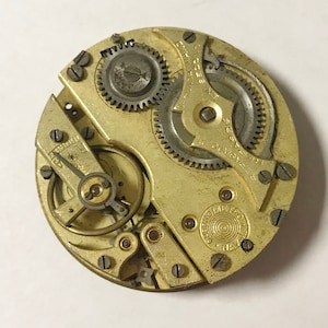 May include: Close-up of a vintage watch movement with intricate gears, screws, and a gold-toned metal finish. The watch mechanism features text that reads "SEGUNDA" and "REPASADO DE". The overall design is circular.