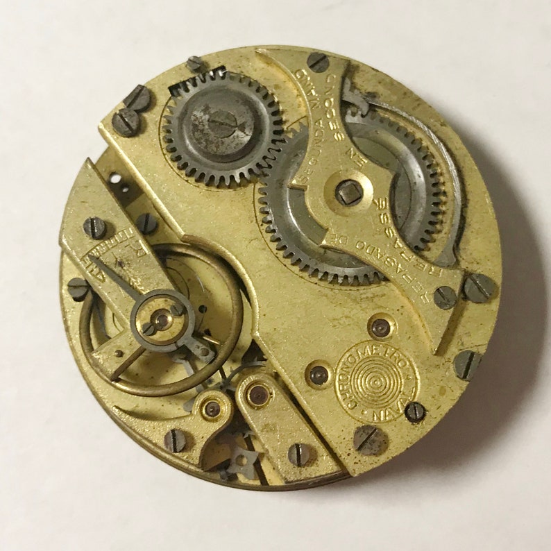 May include: Close-up of a vintage watch movement with intricate gears, screws, and a gold-toned metal finish. The watch mechanism features text that reads "SEGUNDA" and "REPASADO DE". The overall design is circular.