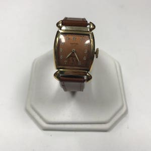 May include: A gold-toned wristwatch with a brown leather band. The watch face is rectangular and has a light brown background with black Roman numeral hour markers. The watch face also has the word "EMPIRE" printed in black.