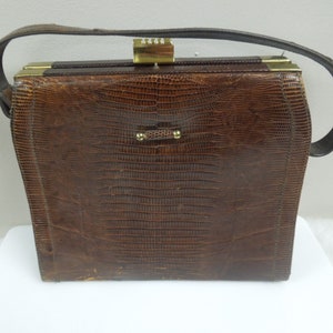 May include: A vintage brown handbag with a textured, reptile-like pattern. The bag features a rectangular shape, a top handle, and gold-toned hardware on the clasp and handle attachments. The bag is likely made of leather.