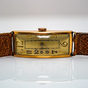 May include: A gold-toned wristwatch with a brown leather band. The watch face has Roman numeral hour markers and Arabic numeral minute markers.