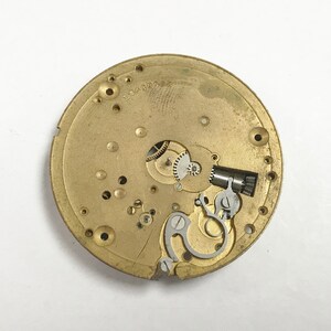K&ouml;nnte beinhalten: Nahaufnahme eines goldfarbenen Uhrwerks mit komplizierten Zahnr&auml;dern und einer silbernen Aufzugsfeder. Das Uhrwerk ist mit "L 0668838" beschriftet.