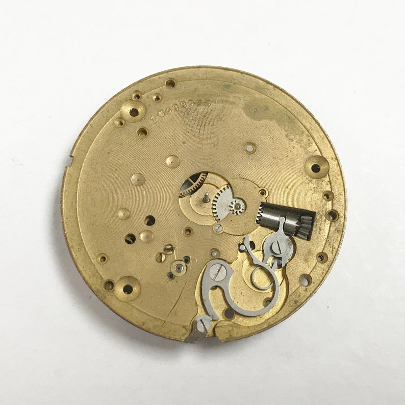 K&ouml;nnte beinhalten: Nahaufnahme eines goldfarbenen Uhrwerks mit komplizierten Zahnr&auml;dern und einer silbernen Aufzugsfeder. Das Uhrwerk ist mit "L 0668838" beschriftet.