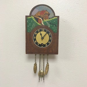May include: A wooden cuckoo clock with a painted scene of a house and a water wheel. The clock face has black Roman numerals and a black hour and minute hand. The clock has a pendulum with three gold-colored weights.