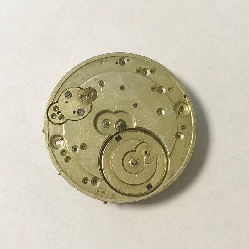 Puede incluir: Primer plano de un movimiento de reloj de tono dorado con engranajes y marcas intrincadas. El movimiento es circular y tiene un di&aacute;metro de aproximadamente 5 cm.