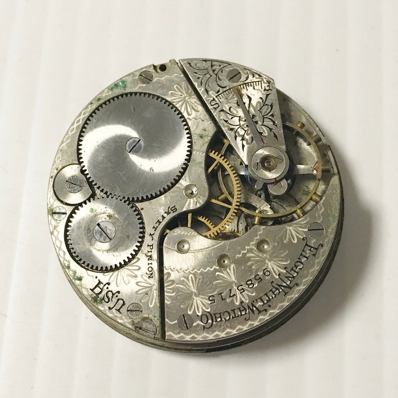 May include: Close-up view of the inner workings of a silver pocket watch. The watch face is engraved with the text "Elgin National Watch Co. 9148886". The watch movement is visible, with gears and a winding stem.