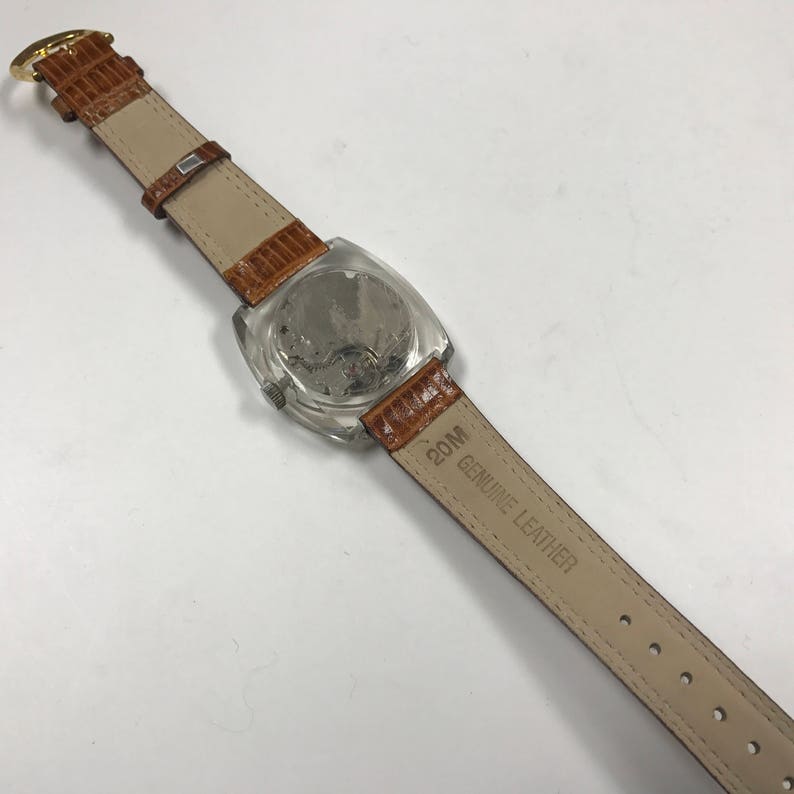 Peut inclure: Un bracelet de montre en cuir marron avec un cadran de montre transparent et le texte "20M GENUINE LEATHER" imprim&eacute; sur le bracelet.
