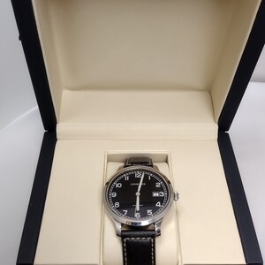 May include: A black-faced wristwatch with silver hands and markers in a dark wood box with the Longines logo. The watch has a black leather band.