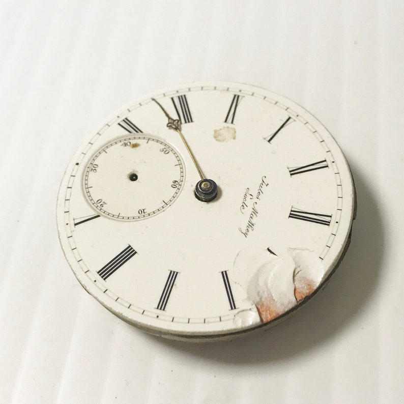May include: A white pocket watch face with black Roman numeral hour markers and a smaller white sub-dial with black markings. The watch face is cracked and chipped. The text "J. Walter & Co.  London" is printed on the watch face.