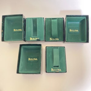 May include: Six Bulova watch boxes. The boxes are rectangular with a dark green velvet interior and a silver-colored exterior. The Bulova logo is printed in gold on the inside of each box.