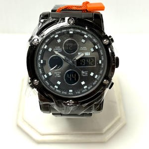 May include: A black analog-digital wristwatch with a round face, featuring a world time bezel with city codes, multiple sub-dials, and a digital display showing 19:42. The watch has a metallic finish, luminous markers, and an orange tag attached.