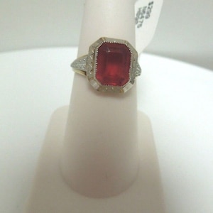 May include: A gold ring with a large, red, rectangular gemstone set in a white metal bezel. The ring is on a white ring display stand.