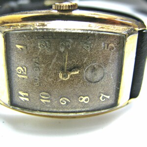 May include: A gold-toned wristwatch with a black leather band. The watch face is rectangular and has Roman numeral hour markers. The watch face is textured and has a small second hand.