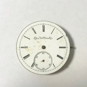 May include: A vintage Elgin National Watch Co. pocket watch face. The white face features black Roman numerals, a smaller seconds dial, and the company's name. The watch face has a metal rim and a small stem.