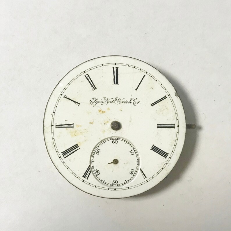 May include: A vintage Elgin National Watch Co. pocket watch face. The white face features black Roman numerals, a smaller seconds dial, and the company's name. The watch face has a metal rim and a small stem.