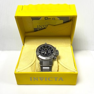 May include: A silver-toned Invicta watch with a black face is displayed in a yellow box. The watch has a stainless steel band and a round face with multiple dials. The box is yellow with the Invicta logo printed on the front.