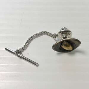 May include: A silver tie tack with a chain and a black oval-shaped top with a gold-colored design.