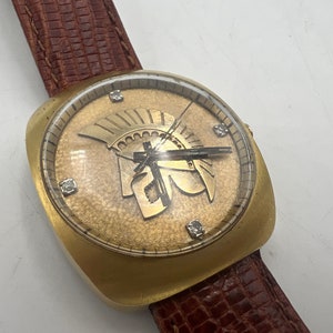 Puede incluir: Un reloj de pulsera dorado con una correa de cuero marr&oacute;n. La esfera del reloj presenta una esfera dorada con n&uacute;meros romanos y un dise&ntilde;o de casco de gladiador dorado.