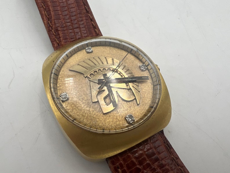 Puede incluir: Un reloj de pulsera dorado con una correa de cuero marr&oacute;n. La esfera del reloj presenta una esfera dorada con n&uacute;meros romanos y un dise&ntilde;o de casco de gladiador dorado.