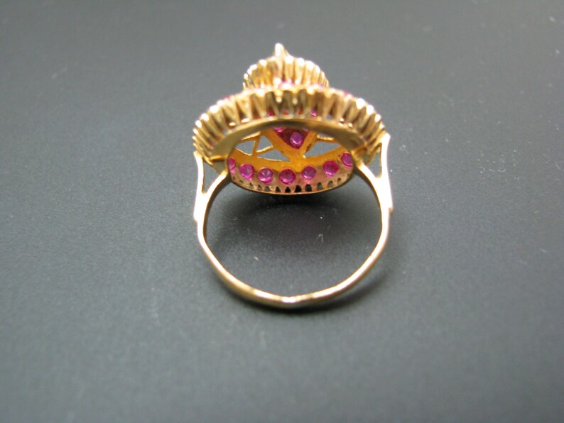 May include: A gold ring with a round, red gemstone surrounded by smaller red gemstones. The ring has a delicate, intricate design.