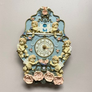 May include: A blue and white clock with cherub figurines and floral accents. The clock face has Roman numerals and a white background. The clock is surrounded by cherubs holding flowers.