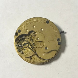 May include: A gold-colored pocket watch movement with the inscription "Elgin National Watch Co." and the number "4194058". The movement has a safety pinion and visible gears.