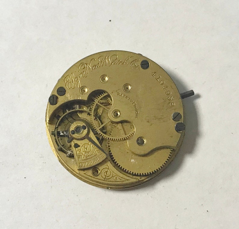 May include: A gold-colored pocket watch movement with the inscription "Elgin National Watch Co." and the number "4194058". The movement has a safety pinion and visible gears.