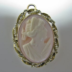 May include: A gold pendant with a cameo featuring a woman's profile. The cameo is set in a pink shell and surrounded by a border of small pearls.