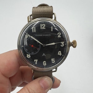 May include: A silver wristwatch with a black face and white numerals. The watch has a brown canvas strap and the words "Ulysse Nardin" printed on the face.