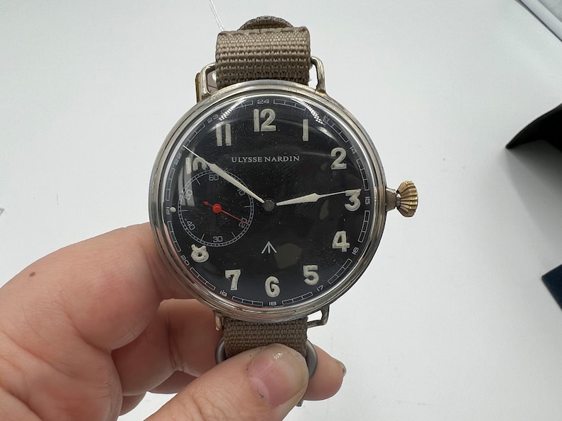 May include: A silver wristwatch with a black face and white numerals. The watch has a brown canvas strap and the words "Ulysse Nardin" printed on the face.
